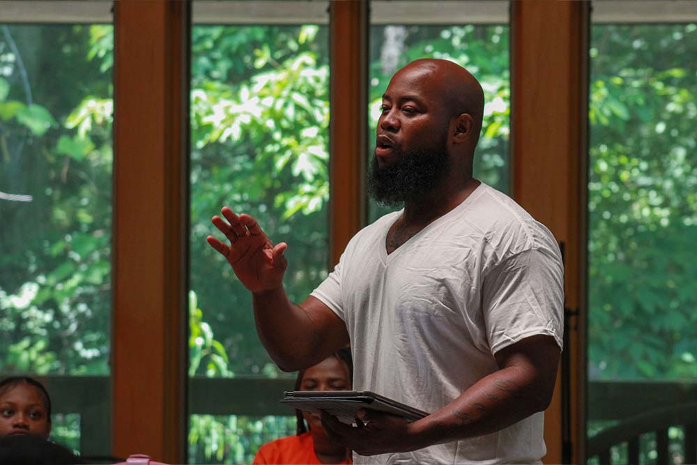 Urban Alliance staff regularly work with the people of our community – here a staff member talks to a group in a room with floor to ceiling windows with green trees outside. It is a peaceful setting.